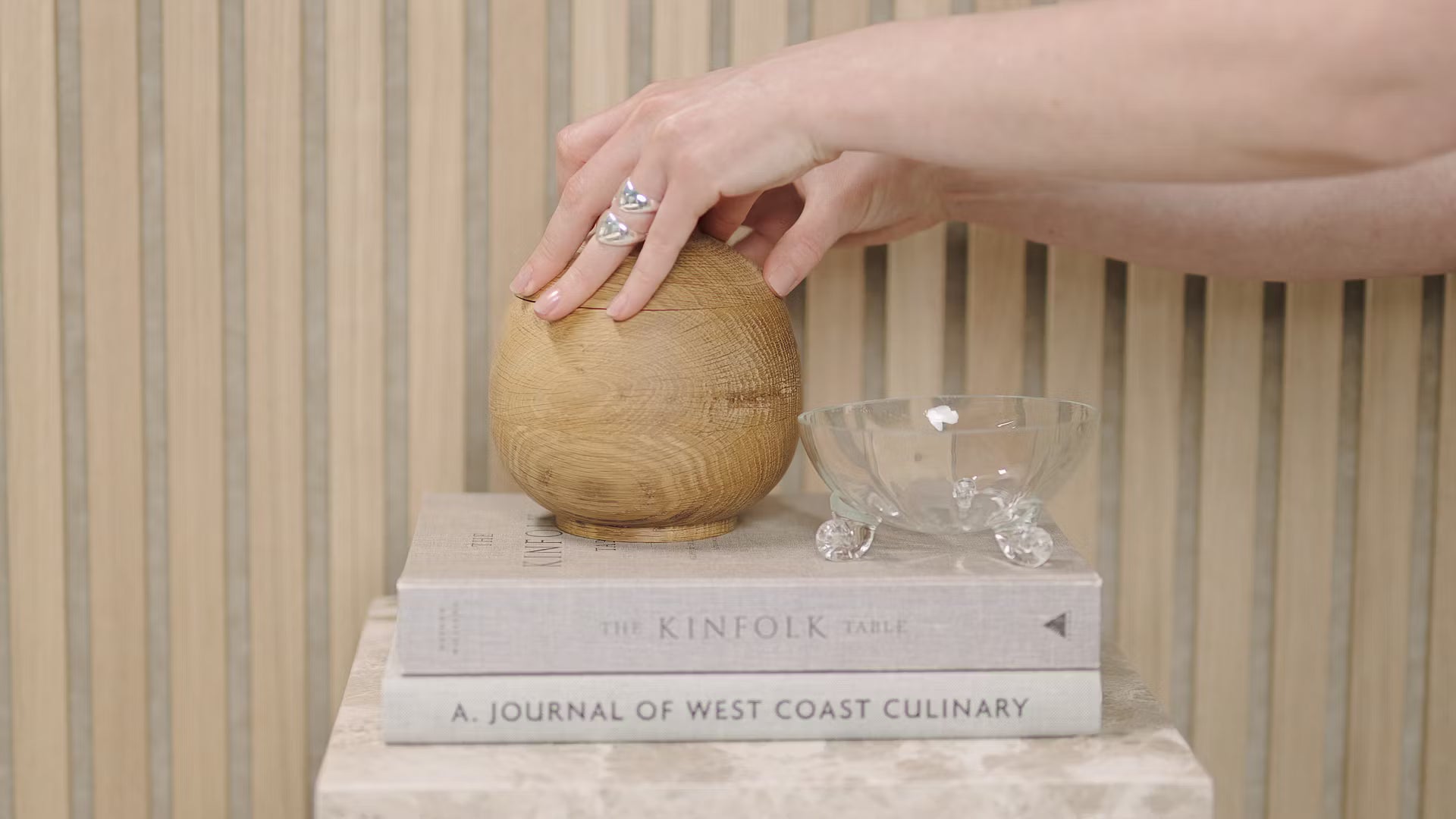 A hand touches a wooden sphere placed on stacked books, including Kinfolk Table and Journal of West Coast Culinary, beside a glass bowl on a wood slat acoustic panel.